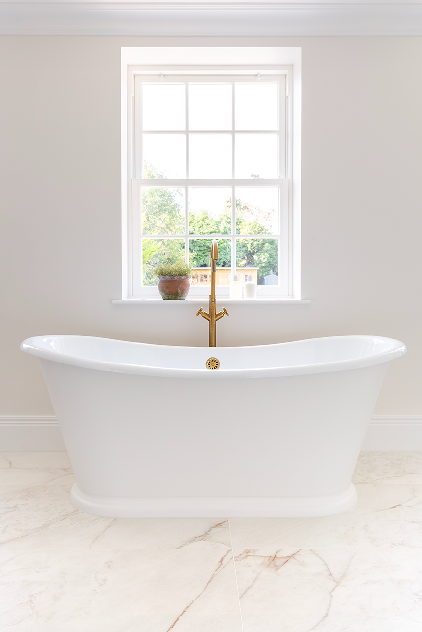 A large freestanding white bath placed infront of a large window with a white marbled floor.