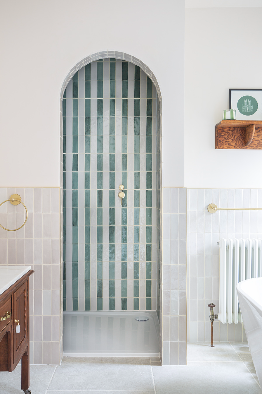 Arched shower niche with Pastello vertical green and white tiles, gold fixtures, and surrounding light gray walls. Nearby are a wooden vanity and towel rack.
