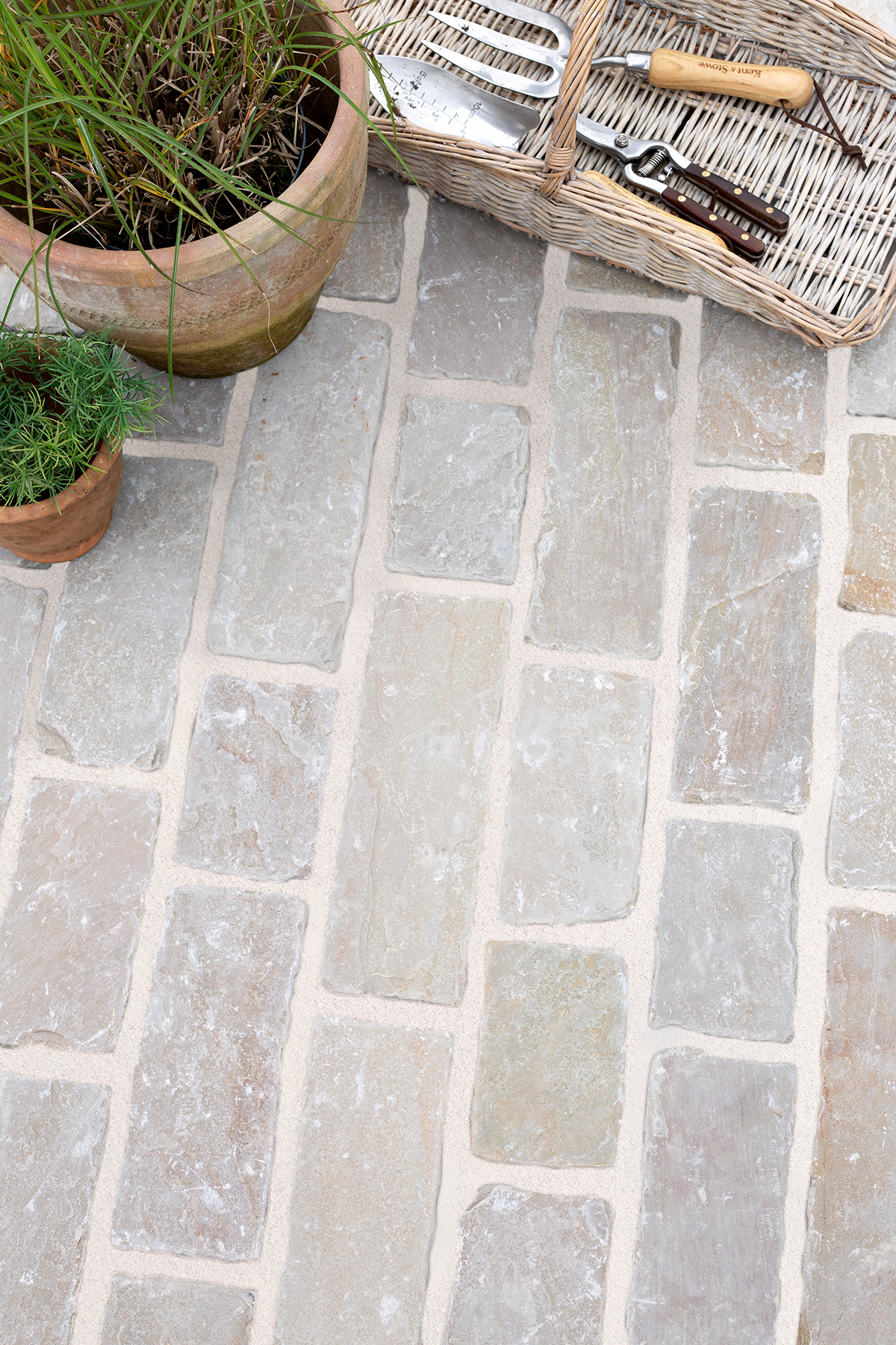 Thistleton Tumbled Sandstone patio with beige and gray bricks. A wicker basket with garden tools sits on the right, flanked by potted plants on the left, creating a rustic feel.