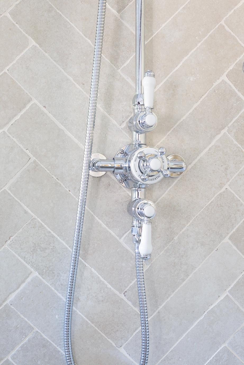 Chrome shower control system against a beige Alnwick Ivory brick herringbone tiled wall. Features three white-handled knobs, a central temperature gauge, and sleek hose.