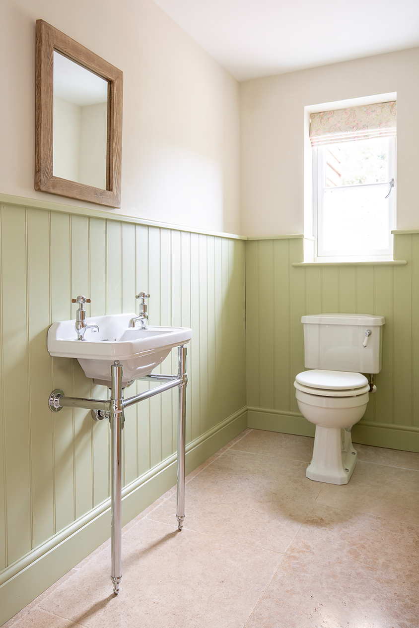 A sage green bathroom with a white toilet and sink, a beige tiled floor and a wooden mirror on the wall.