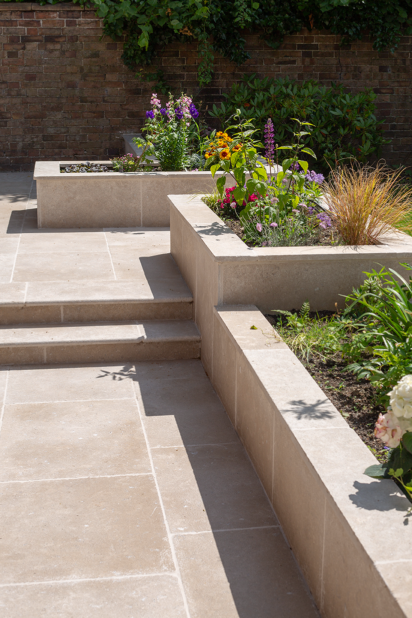 Terraced garden with Dijon Seasoned Limestone steps and retaining walls, featuring blooming flowers like lavender and sunflowers under bright sunlight. Brick wall backdrop.