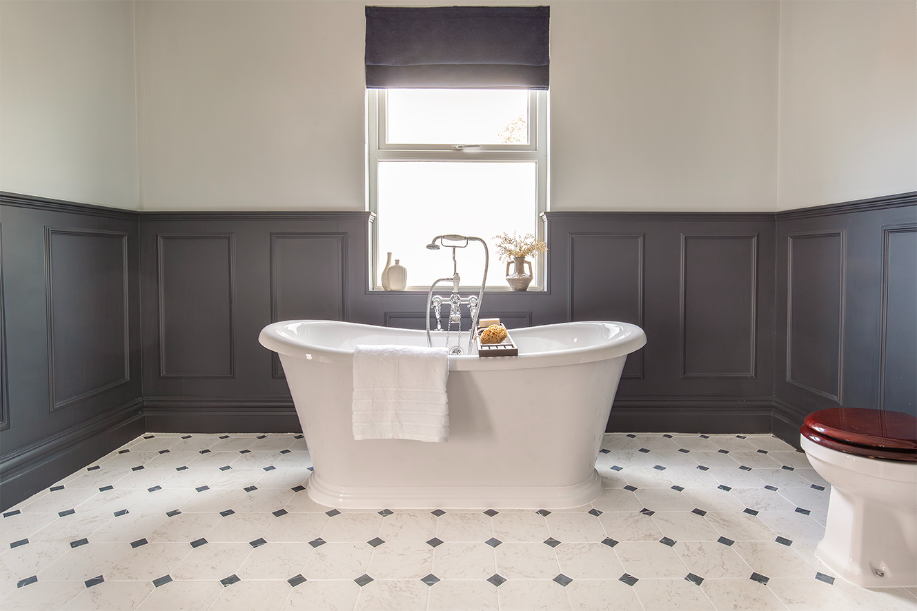 A large bathroom with a white freestanding bath and toilet, with a limestone tiled floor and dark panelling..