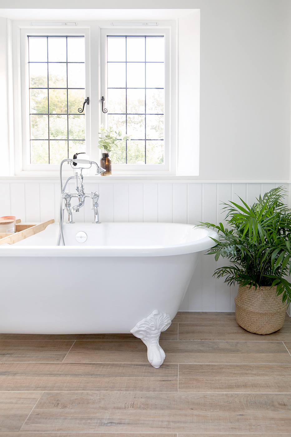 Bright bathroom with a classic white clawfoot tub, wooden tray, and silver faucet. Sunlit window, white paneling, potted plant, and Henbury Natural Oak porcelain wood floor add warmth.