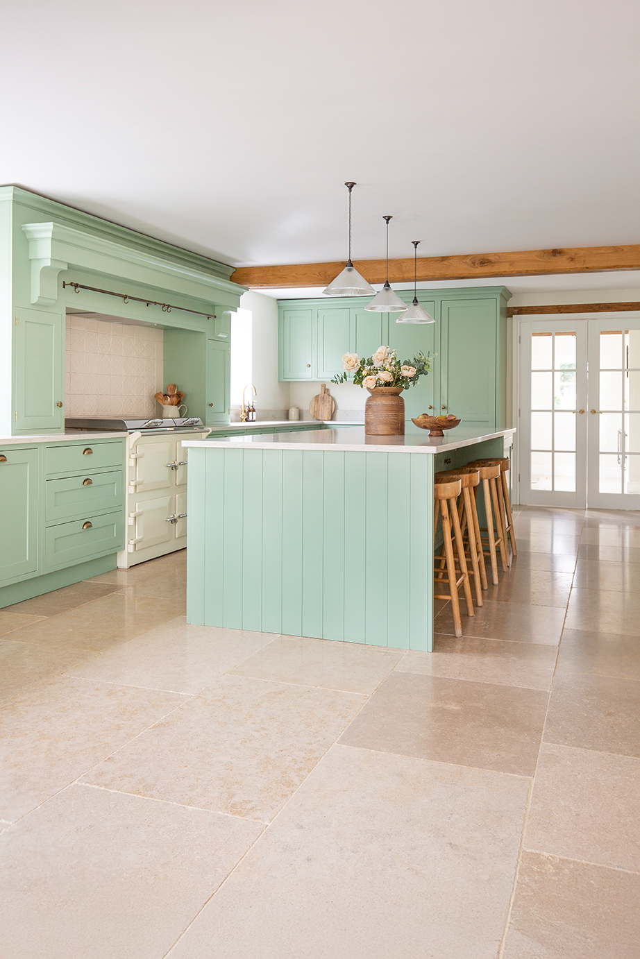 A bright, French inspired green kitchen with a large island and wooden bar stools and a limestone floor.