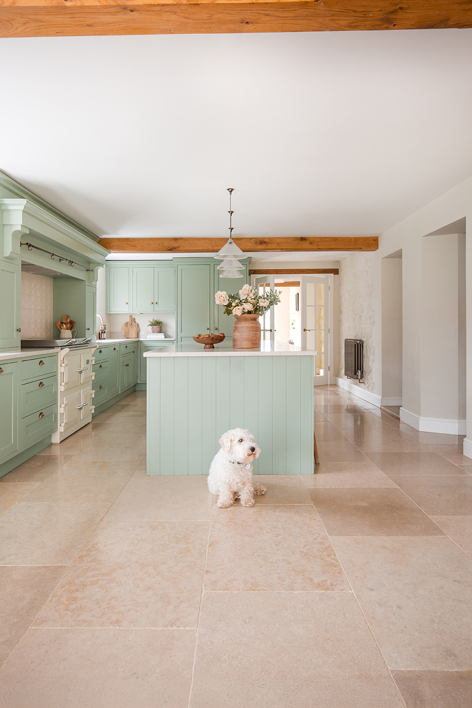 A bright, French inspired kitchen with a white cooker and a beige limestone tiled floor. A white small dog sits on the limestone tiles.