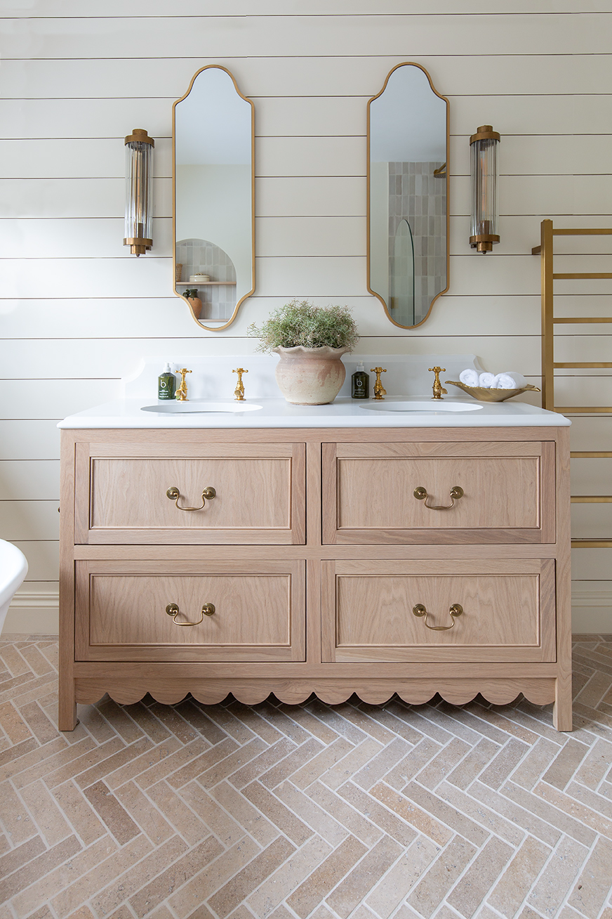 Elegant bathroom with double wooden vanity, gold fixtures, and two mirrors. Potted plant and folded towels add a cozy touch ith Dijon tumbled herringbone floor. Bright, serene ambiance.