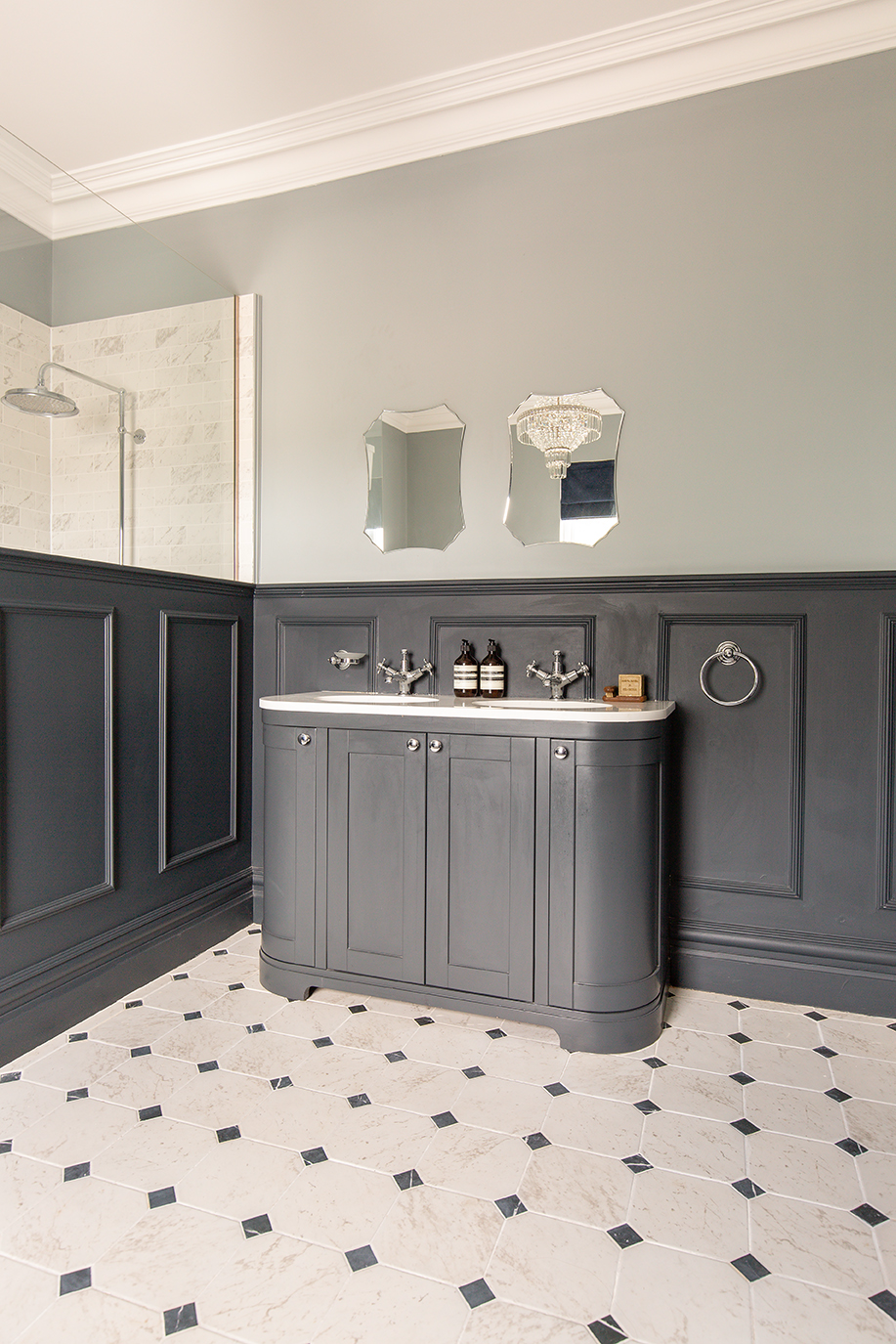 A dark grey bathroom vanity unit with 2 mirrors, dark panelled walls and a limestone tile floor.