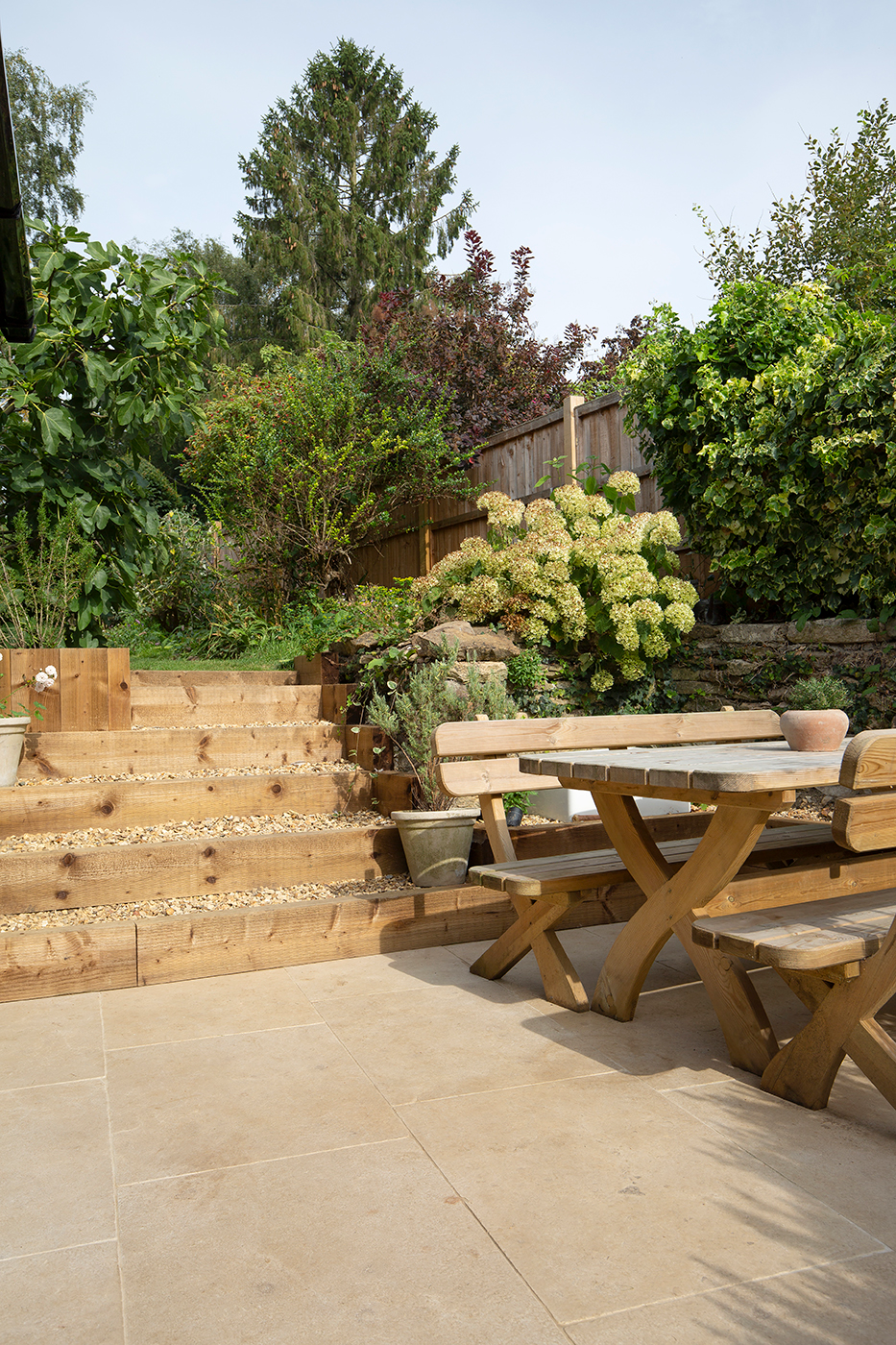 Sunny garden Tuscany Seasoned Limestone patio with wooden table and benches. Wooden steps lead up to lush greenery and flowers. Peaceful, inviting outdoor setting.