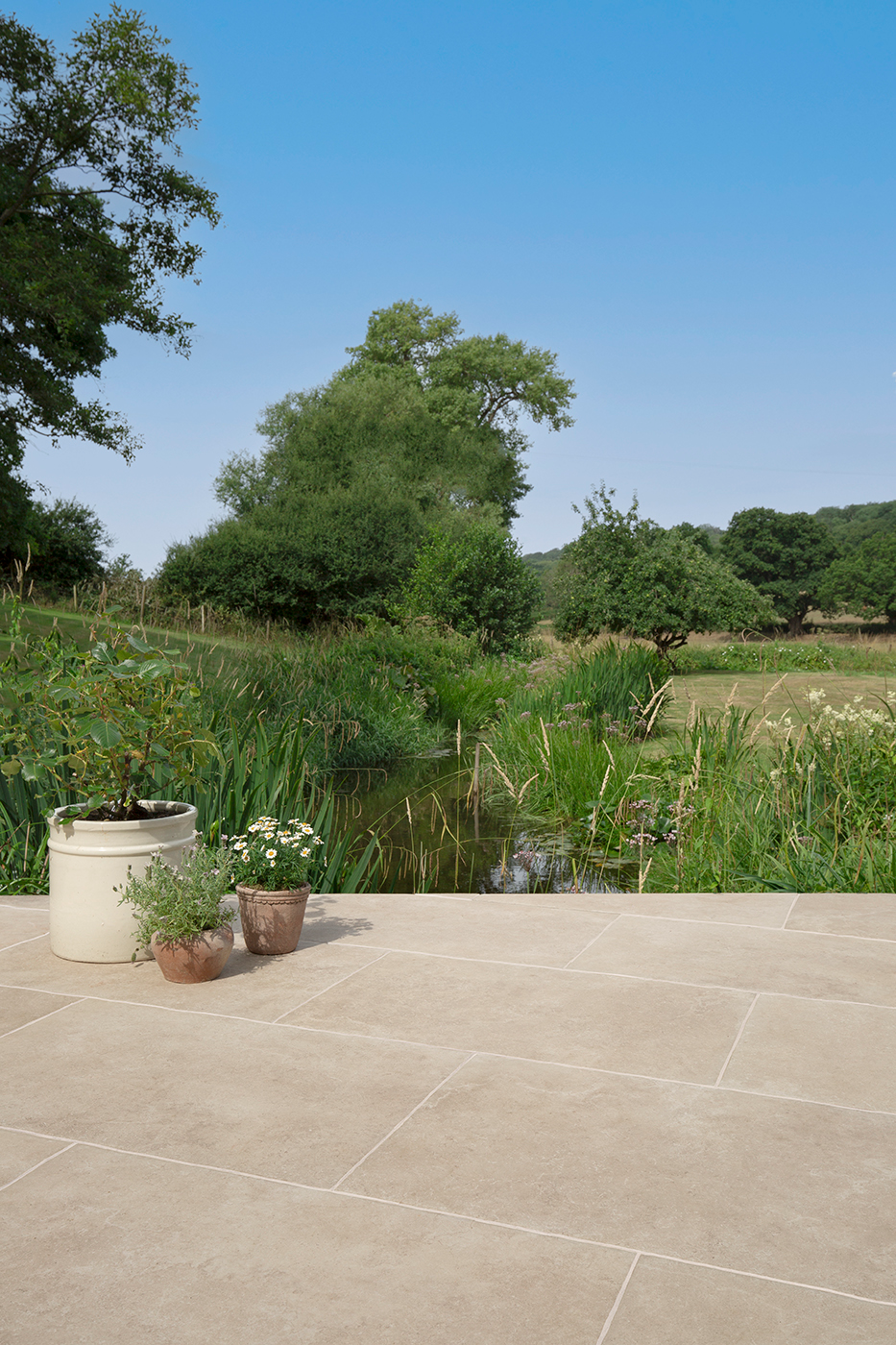 A serene garden scene with three potted plants on a Hambleton Beige Stone Effect tiled patio overlooking a lush pond and greenery under a clear blue sky.