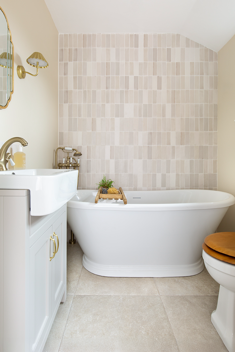 Elegant bathroom with a white freestanding bathtub against a beige tiled wall and Hambleton Ivory floor. A wooden bath tray holds a plant. Light tones create a calm ambiance. tones create a calm ambiance.