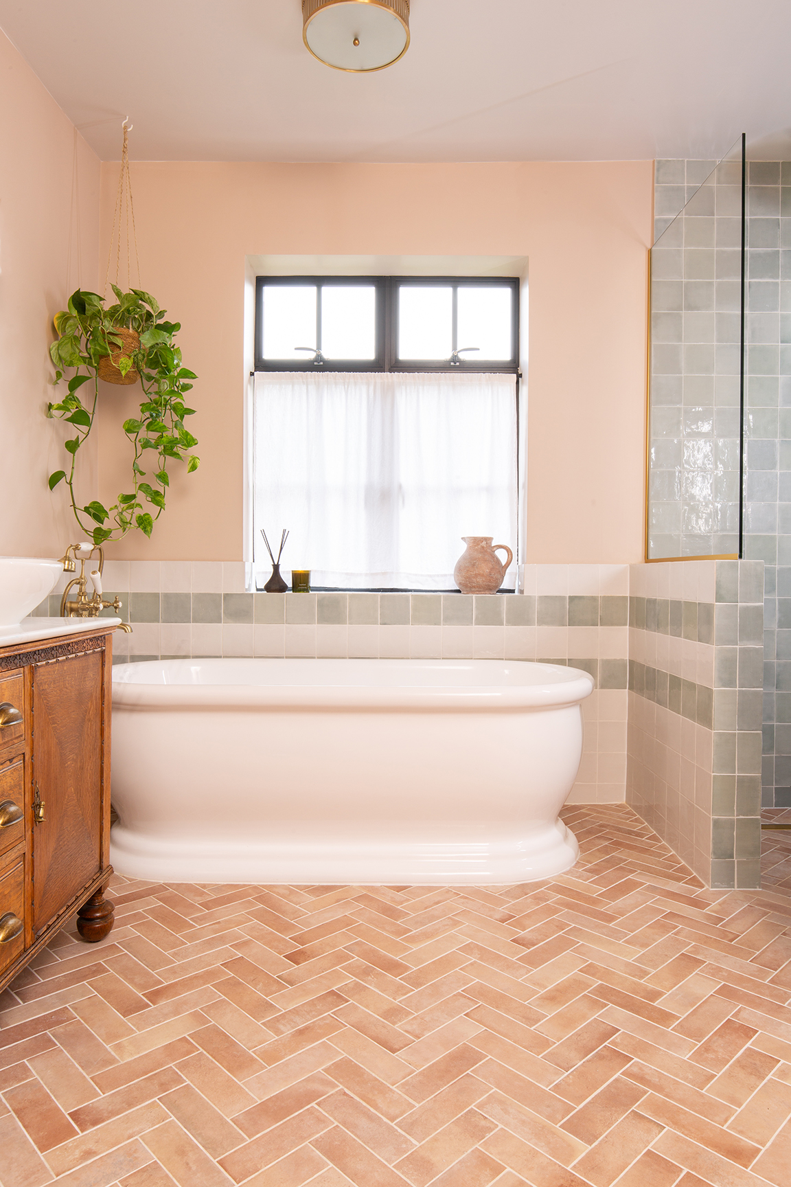 A cozy bathroom with a white freestanding tub against pale green tiles. A window with sheer curtains is above, flanked by plants and pottery. Warm, welcoming tones are set by Campello Rosato herringbone-patterned brick flooring and soft lighting.
