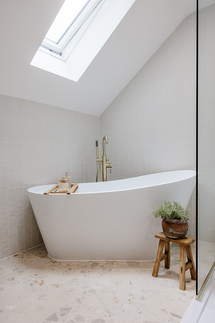 Modern bathroom with a white standalone tub under a skylight. A small wooden stool with a potted plant adds a natural touch on top of Orottelli Avorio flooring. Light tiles and airy ambiance.