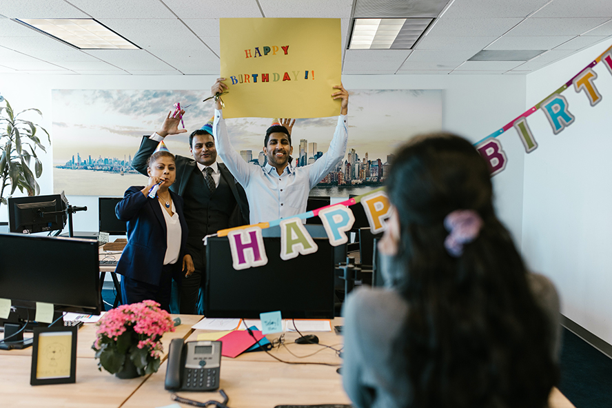 Coworkers holding a sign that reads "happy birthday". They're suprising their coworker.